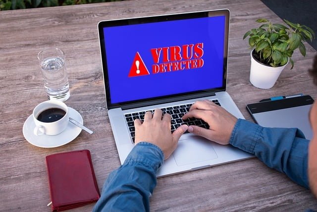 A person working on a laptop with a "Virus Detected" warning displayed on the screen, surrounded by a coffee mug, plant, and book on a wooden desk.